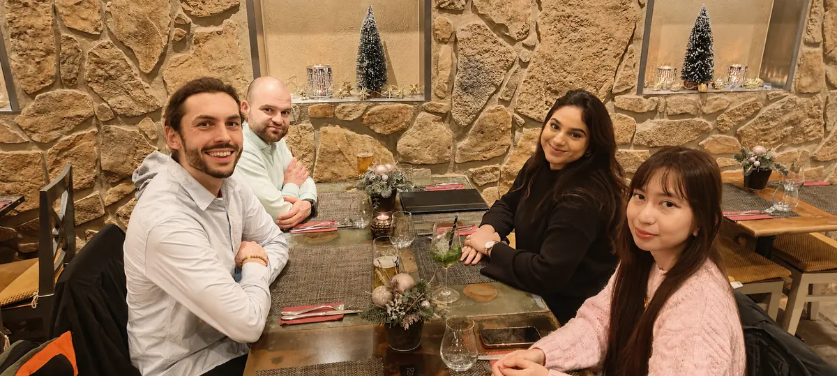 Jan Edinger lacht in die Kamera, an einem gedeckten Tisch in einem Restaurant mit Natursteinwand und weihnachtlicher Dekoration; neben ihm sitzen drei weitere Gäste.
