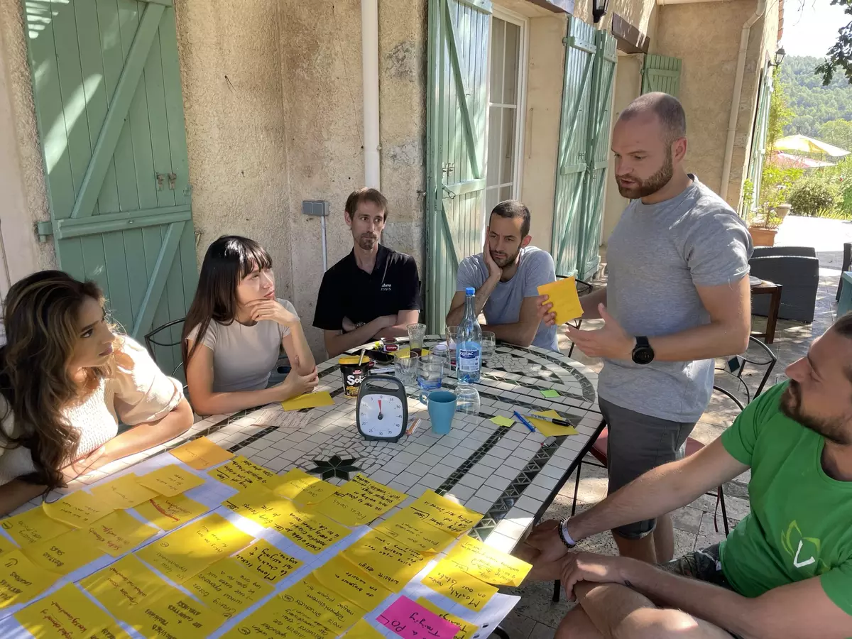 Marwan El Khalil sitzt mit dem Vishnu-Team um einen Mosaiktisch vor einem Steinhaus mit grünen Fensterläden; auf dem Tisch und einem Papierbogen kleben dicht an dicht gelbe Haftnotizen.