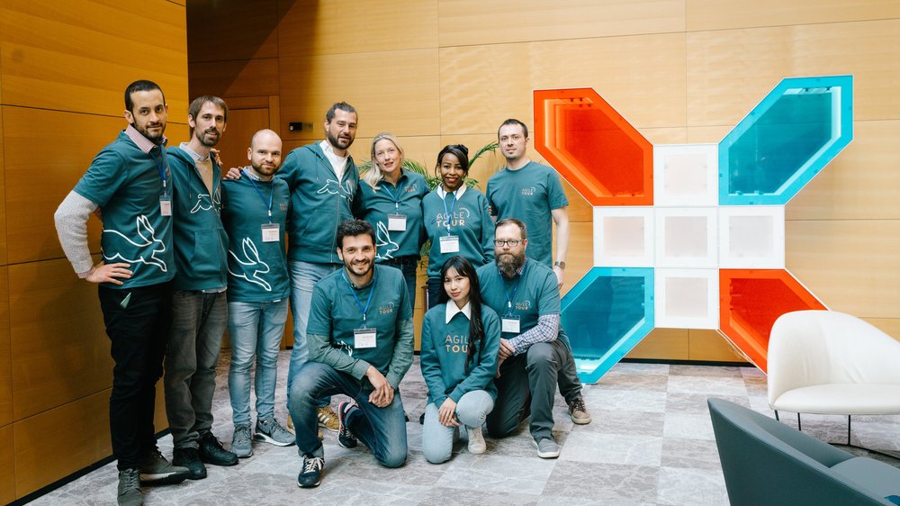 L’équipe d’organisation de l’Agile Tour en maillots bleu-vert près d’une installation lumineuse orange et bleue.