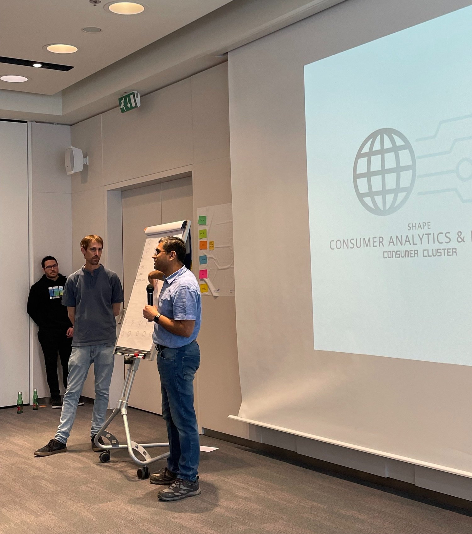 Workshop-Szene: ein Mann spricht ins Mikrofon neben einem Flipchart, an der Wand eine Folie zu Consumer Analytics.