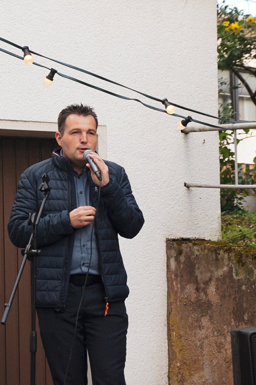 Ein Mann spricht bei der Eröffnungsfeier ins Mikrofon, über ihm eine Lichterkette.