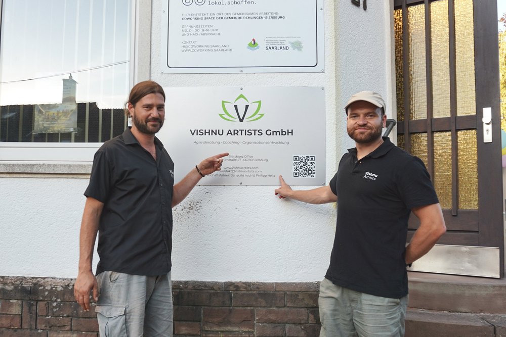 Deux hommes désignent la plaque de Vishnu Artists GmbH à côté de l’entrée.