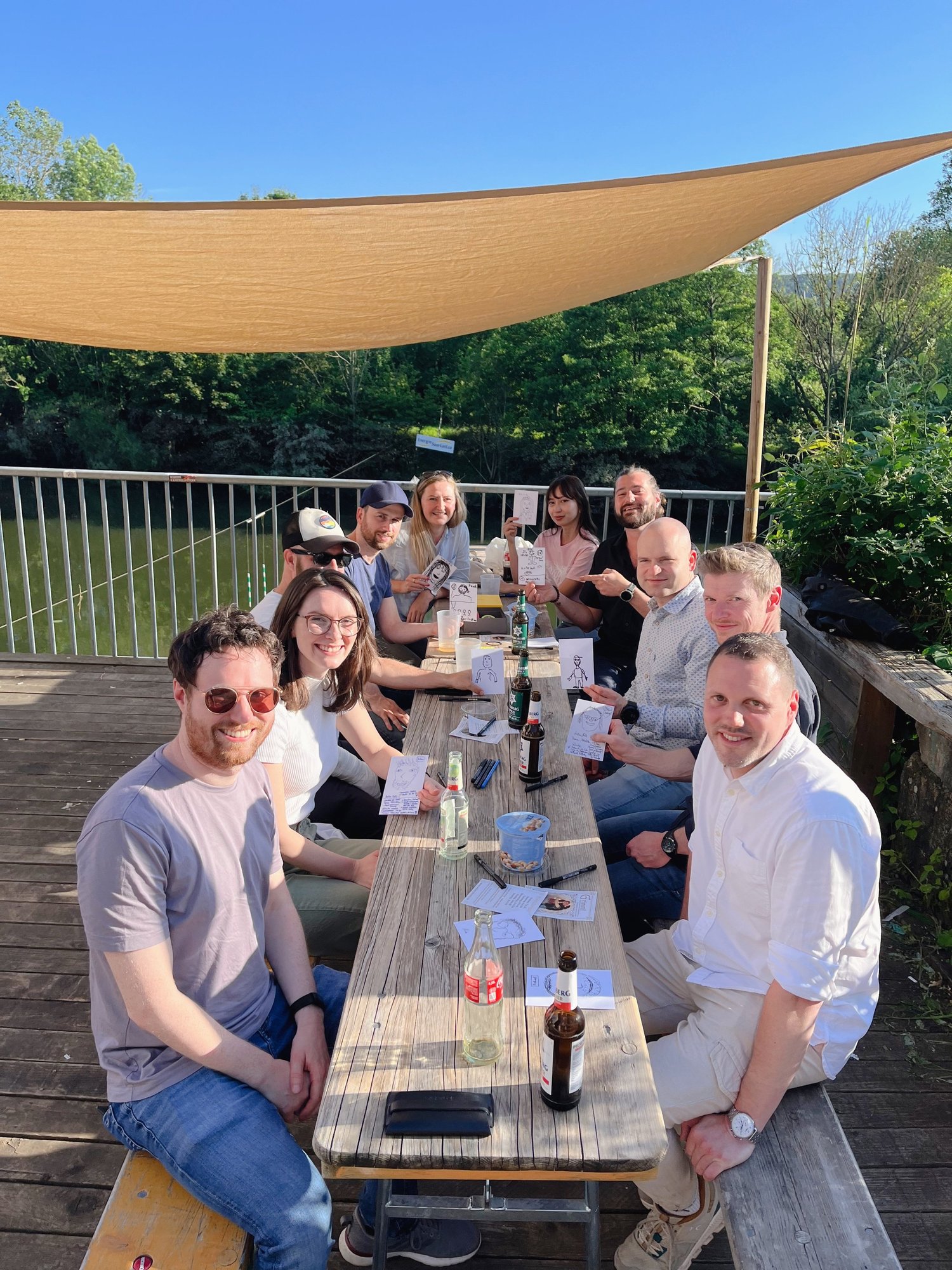 Meetup autour d’une longue table en bois sur un ponton au bord de l’eau, les participants montrent des portraits dessinés.